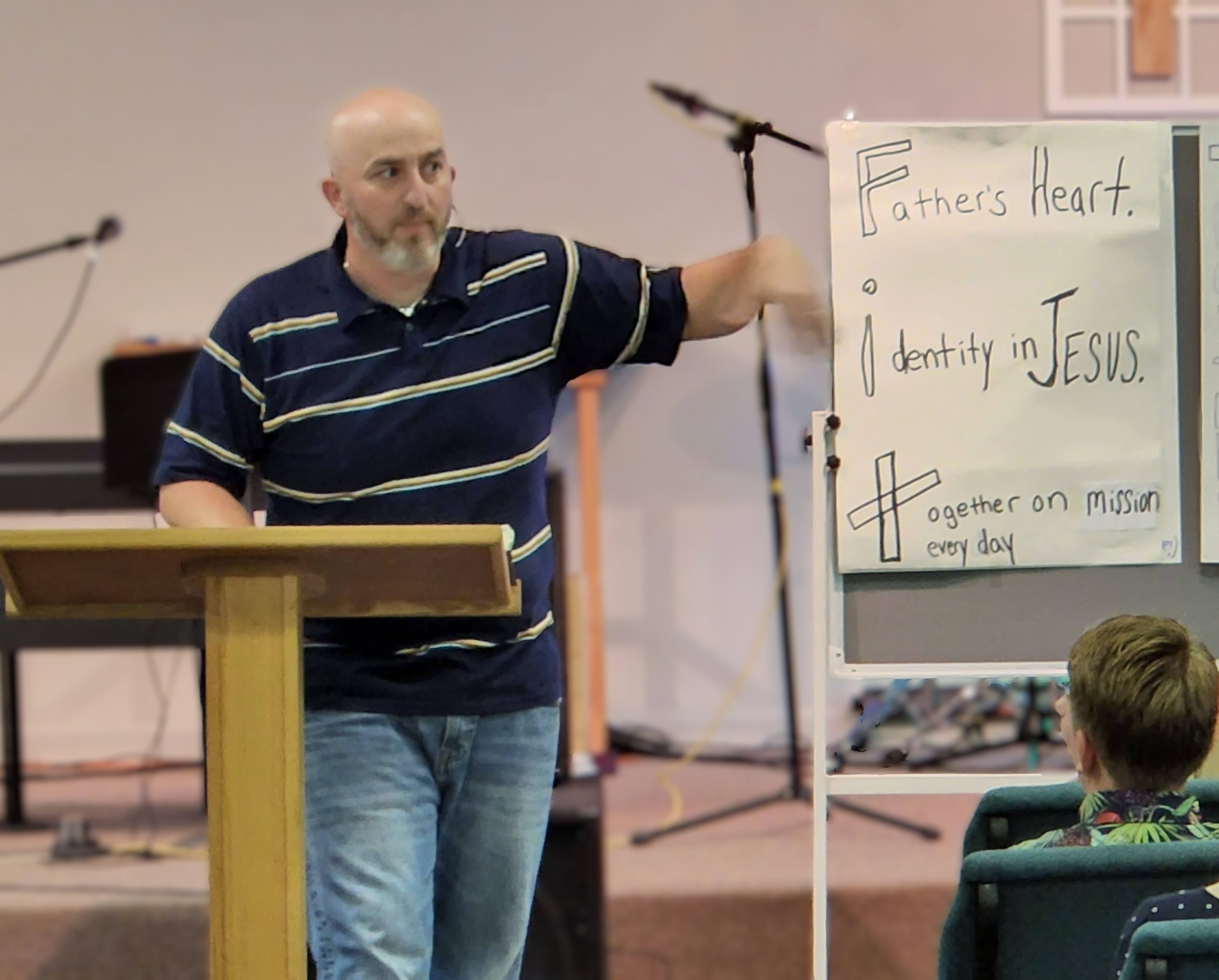 Pastor preaching at chapel service. A flip chart shows the acronym FIT: Father's heart; Identity in Christ; Together on mission.